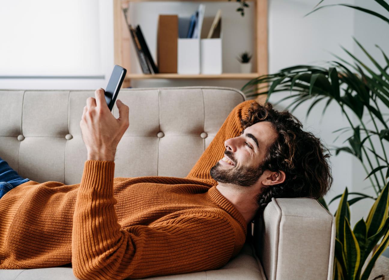 Junger Mann liegt entspannt auf einem Sofa und schaut in sein Handy